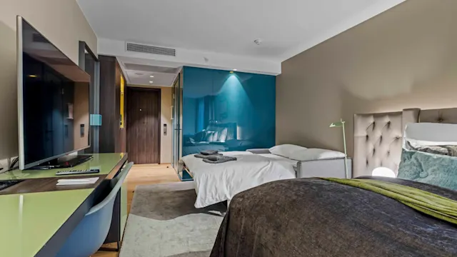Et moderne hotellrom med to pent oppredde senger. Et skrivebord med stol og stor TV er til venstre. Rommet har en grønn aksentvegg og varm belysning.