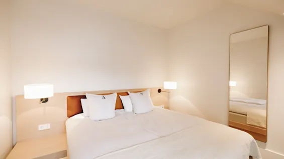Ljust och minimalistiskt hotellrum med en dubbelsäng, varma sänglampor och en helkroppsspegel.
