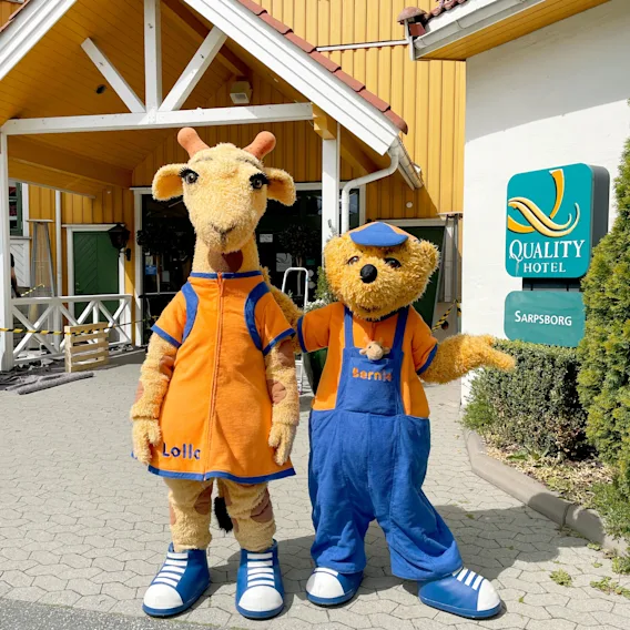 To maskoter, en sjiraff og en bjørn, står utenfor et Quality Hotel i Sarpsborg og ønsker gjester velkommen.