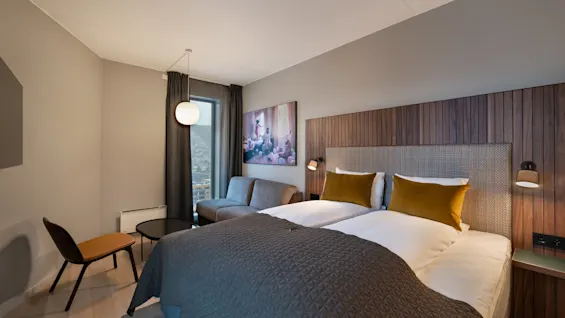 En pent oppredd seng med sennepsgule puter står under ambient belysning ved siden av en moderne vegg. I nærheten er en stol og et bord plassert ved et vindu med mørke gardiner.