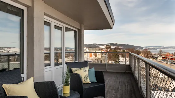 A balcony features two black wicker chairs with yellow and patterned cushions, next to a yellow potted plant. Overlooks a snowy urban landscape under a partly cloudy sky.