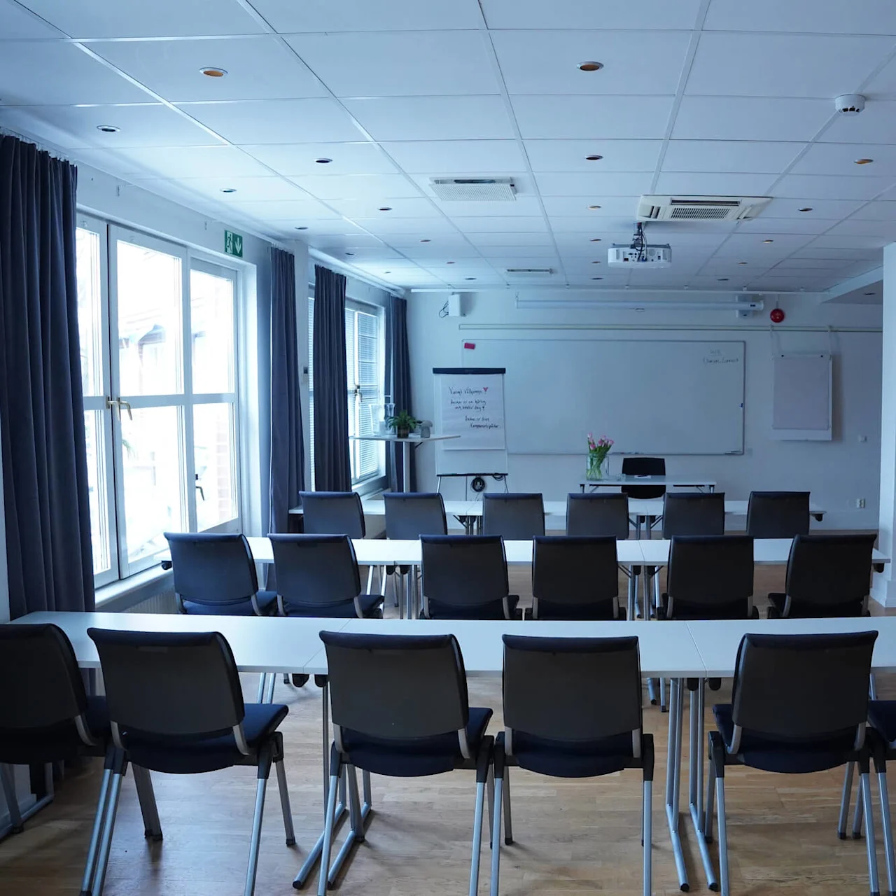 Conference room arranged with tables and chairs facing a whiteboard. Natural light from windows, no people present.