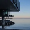 Moderne bygning hænger over roligt hav ved tusmørke, pæle og klipper synlige, fredelig havhorisont.
