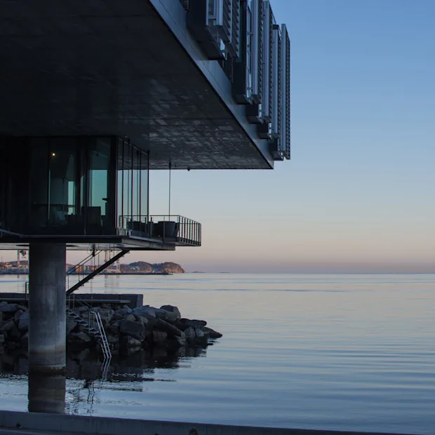 Modern building overhanging calm sea at dusk, pilings and rocks visible, tranquil ocean horizon.