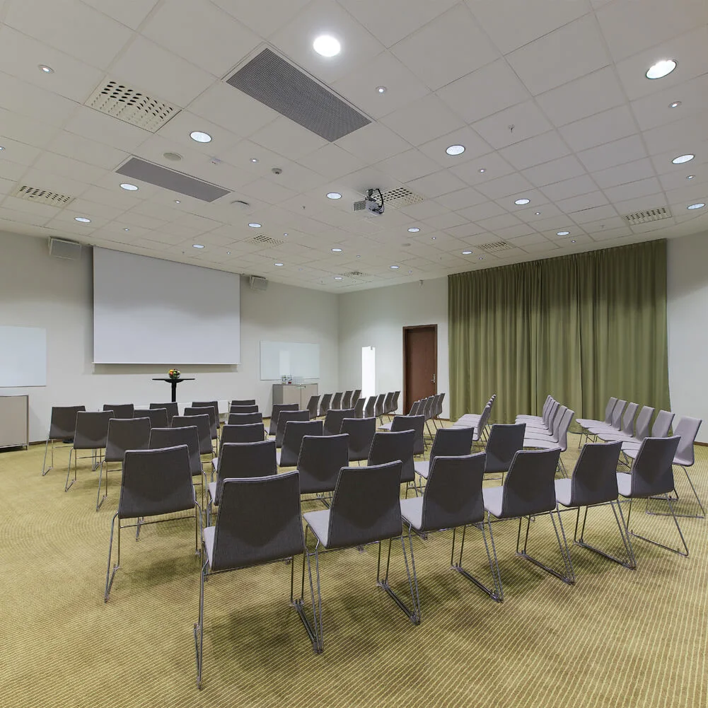 Rows of empty gray chairs face a large white screen in a spacious conference room with green curtains and beige carpet. Projector and speakers are mounted on the ceiling.