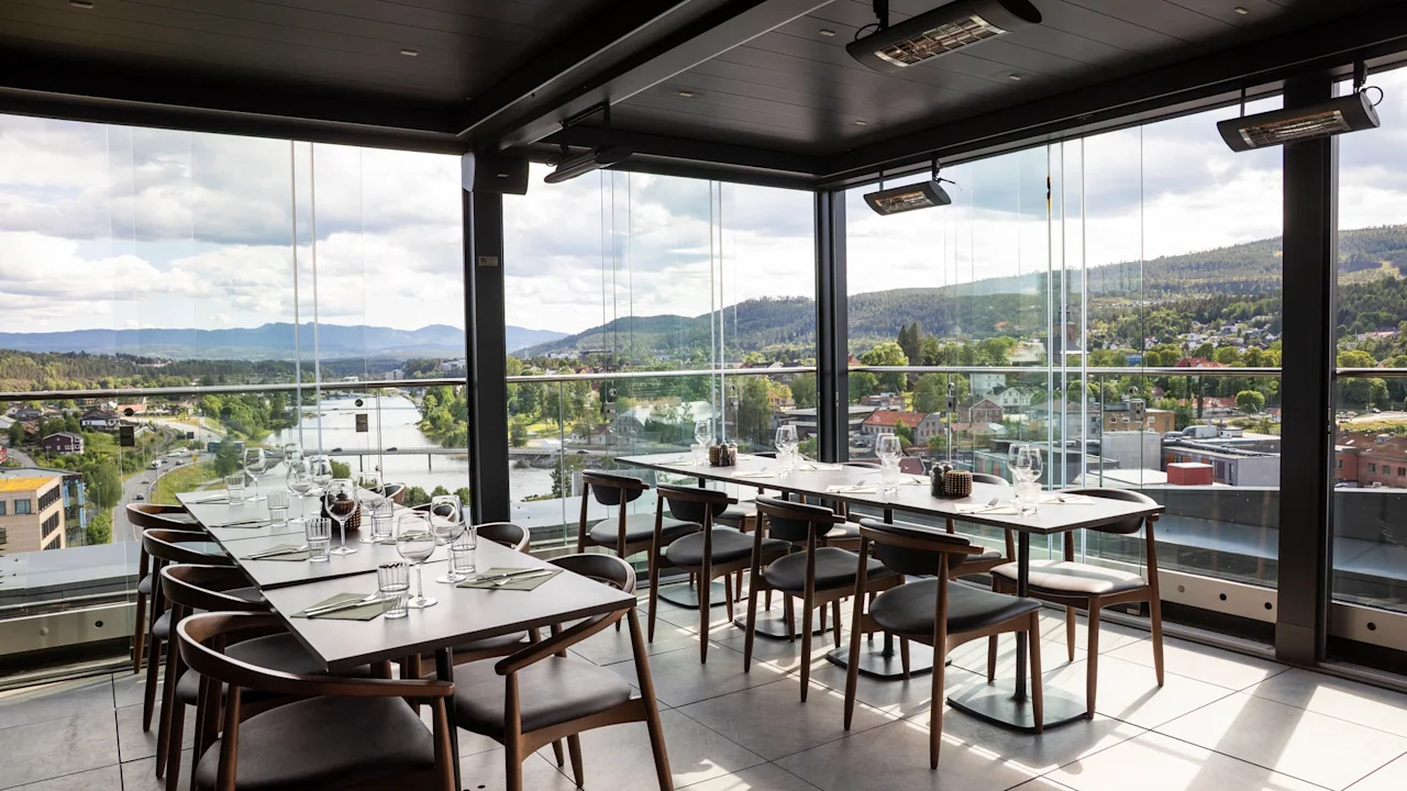 En moderne restaurant med gulv-til-loft-vinduer, der tilbyder en naturskøn udsigt over en by, flod og bjerge. Bordene er dækket til spisning.