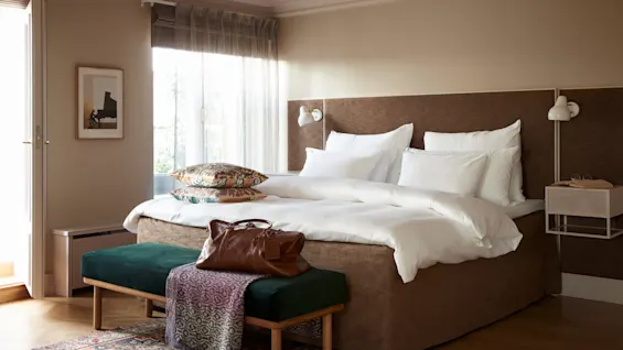 En pent redt seng med hvitt sengetøy og dekorative puter står i et koselig soverom. En brun veske ligger på en grønn benk ved fotenden av sengen.