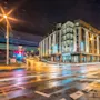 A modern hotel building with illuminated windows stands on a street corner at night, surrounded by light trails from passing vehicles. Text: "Comfort Hotel."