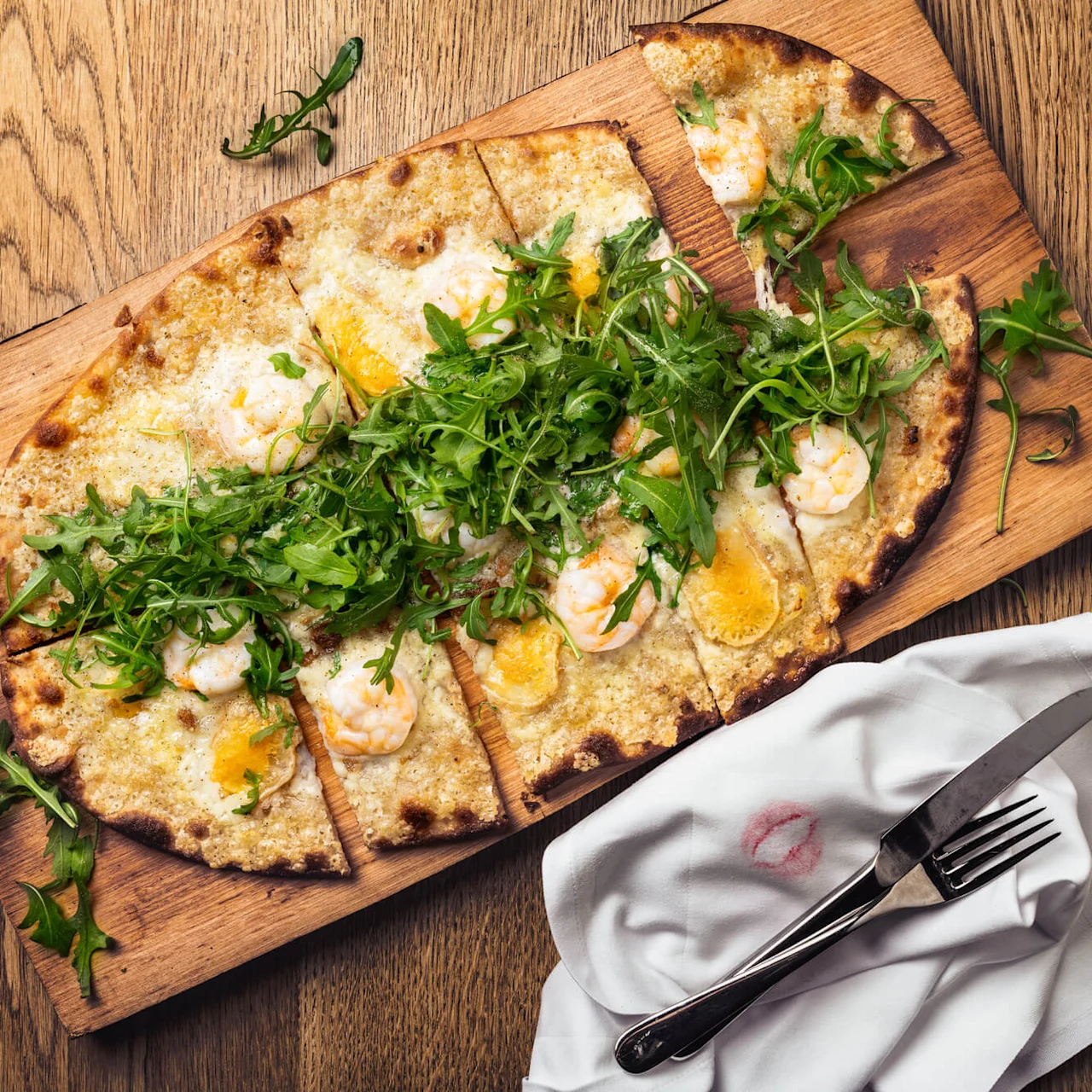 A flatbread pizza topped with arugula and shrimp is sliced on a wooden board. A fork and knife rest on a napkin with a lipstick mark on a wooden table.