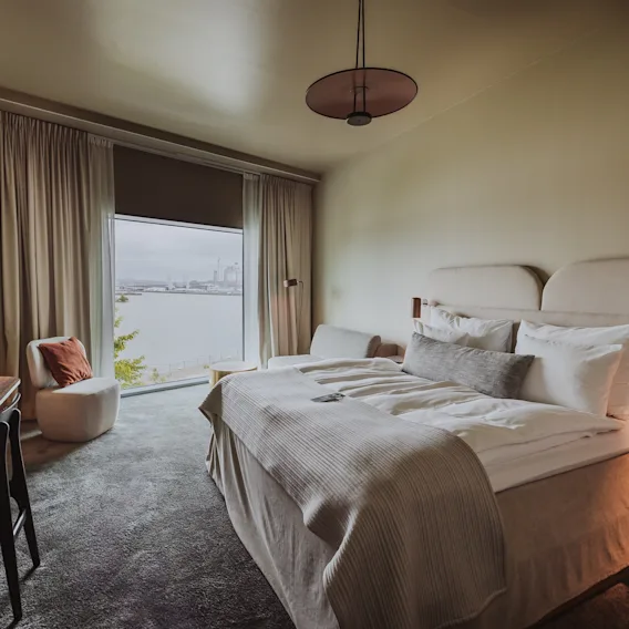 En prydligt bäddad säng med vita sängkläder står i ett modernt hotellrum, med sidobord och lampor. Ett stort fönster visar en naturskön utsikt över vattnet utanför.