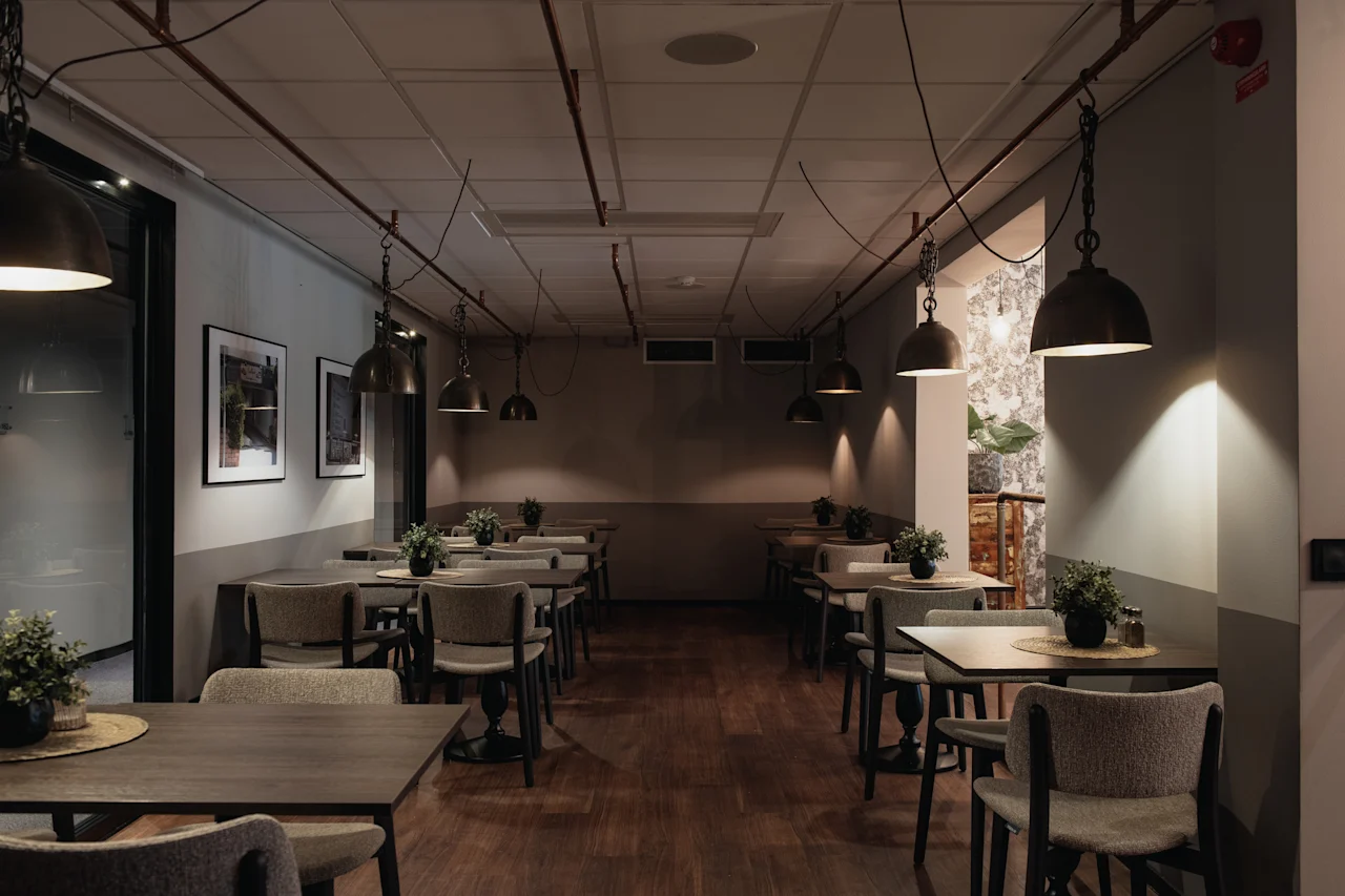 A dimly lit restaurant dining area with multiple tables and chairs, decorated with small plants and framed art on the walls.