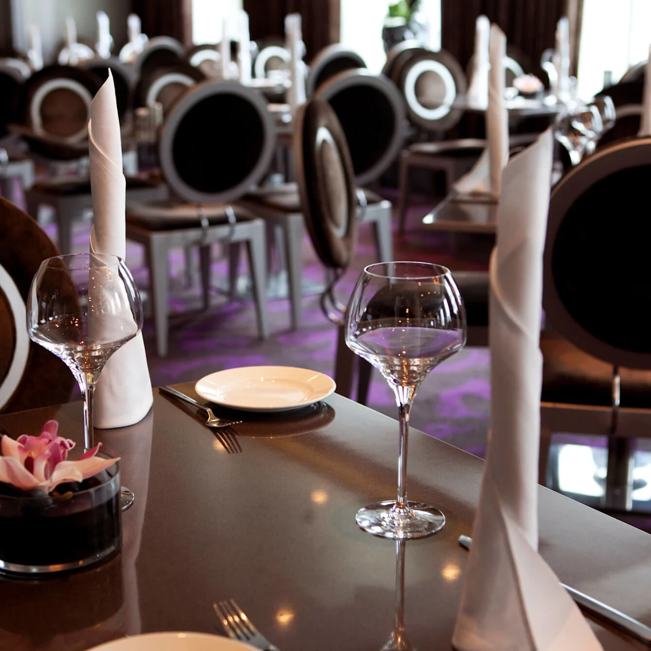 Et restaurantbord er dækket med vinglas, foldede servietter, bestik og en floral dekoration. Baggrunden viser flere elegante spisestole under blød belysning.