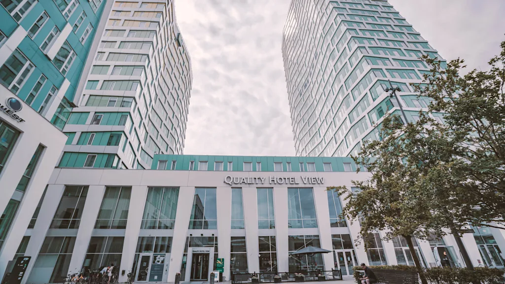 Two tall, modern buildings rise with a connecting structure labeled "Quality Hotel View." Trees and a cloudy sky surround the hotel entrance.
