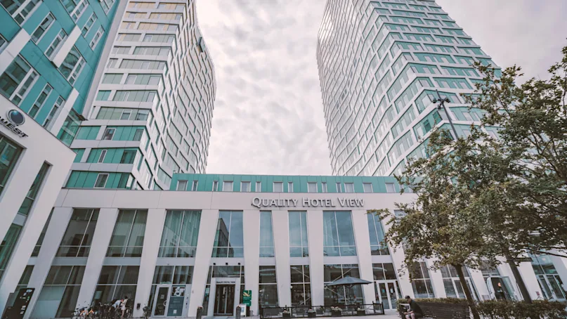 Two tall, modern buildings rise with a connecting structure labeled "Quality Hotel View." Trees and a cloudy sky surround the hotel entrance.