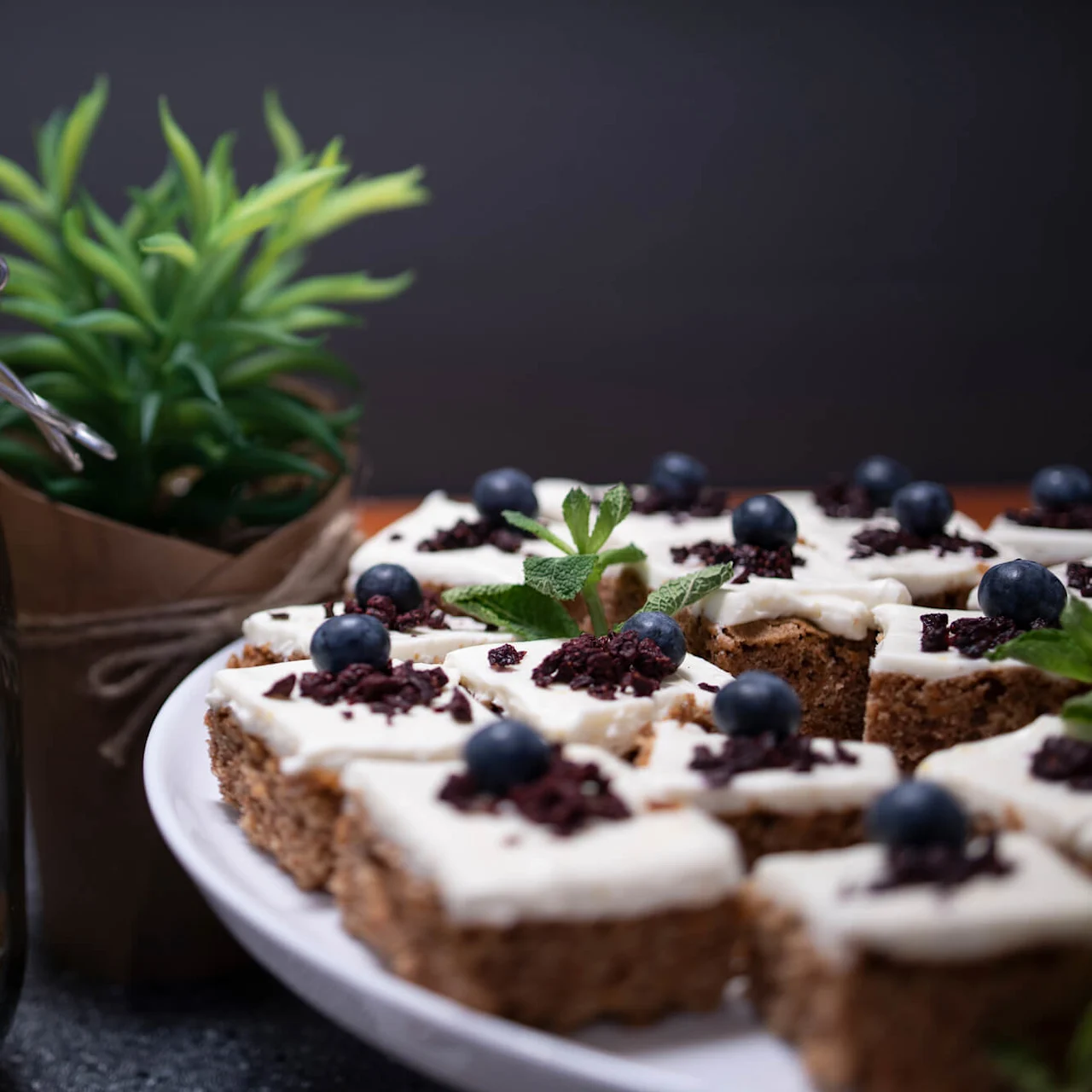 Glasburk innehåller sockerprydda kakor. Vit tallrik har frostade fyrkantiga kakor med blåbär och myntablad. Grön växt finns i bakgrunden, vilket skapar en mysig köksmiljö.