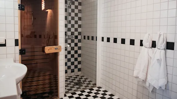 Sauna med en stängd glasdörr med träram, i ett kaklat badrum med rutigt golv och väggar, duschkabin och två vita handdukar på väggen.