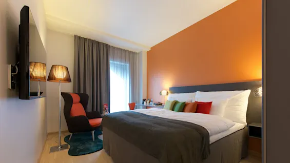 Ett modernt sovrum har en stor säng med färgglada kuddar bredvid en läsfåtölj. En golvlampa lyser upp utrymmet, kompletterat med en orange accentvägg.