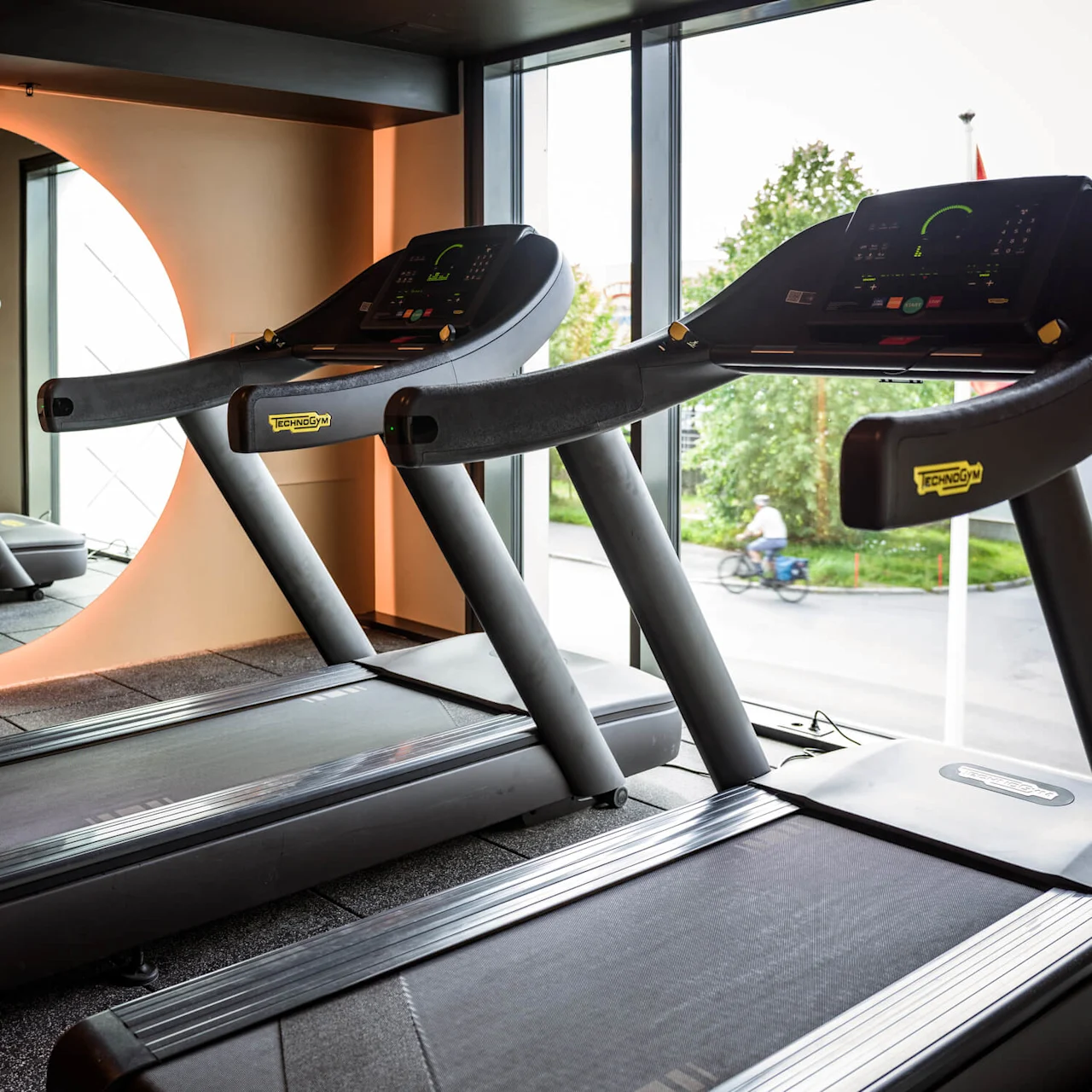 Tredemøller står stille på et treningssenter, vendt mot et stort rundt speil med oransje belysning. Utenfor vinduet sykler en person forbi trær. Merke: TECHNOGYM.