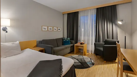 Et pænt arrangeret hotelværelse har en seng, sofa, lænestol og bord med et tæppe på trægulvet og indrammede billeder på væggen. Værelset er varmt oplyst med gardiner.