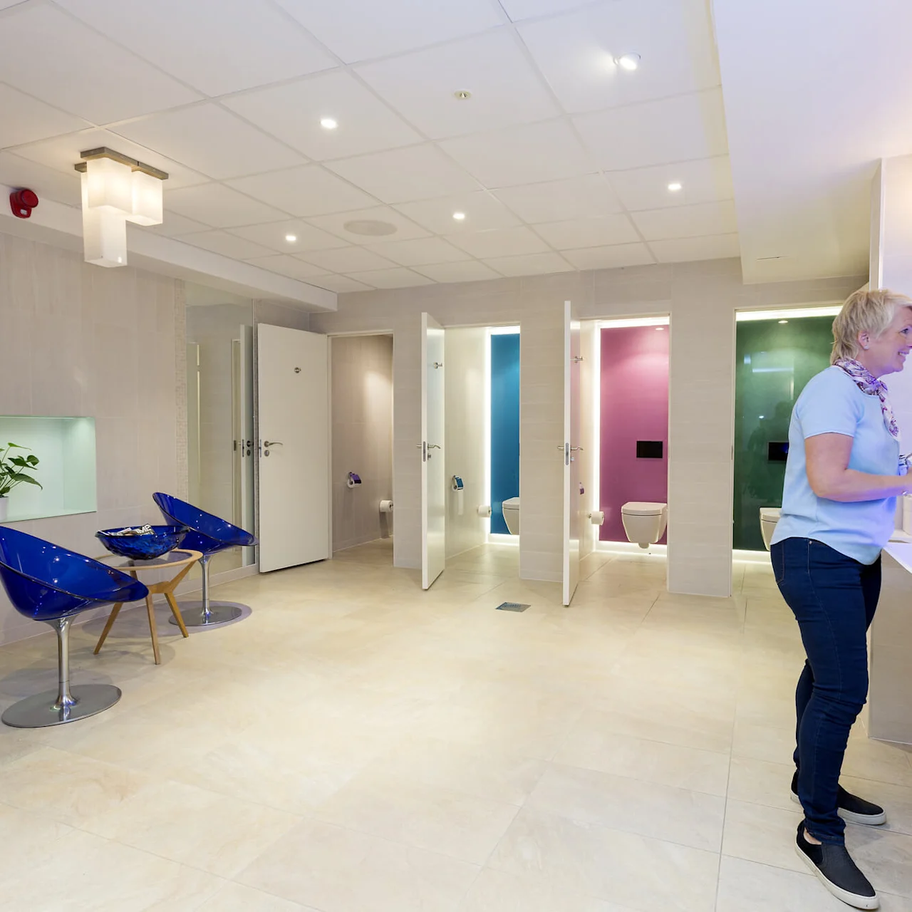 En kvinde vasker hænder i et moderne badrum med farverige toiletbåse og blå stole. Rummet er rent og godt oplyst, med sammenrullede håndklæder på bordet.