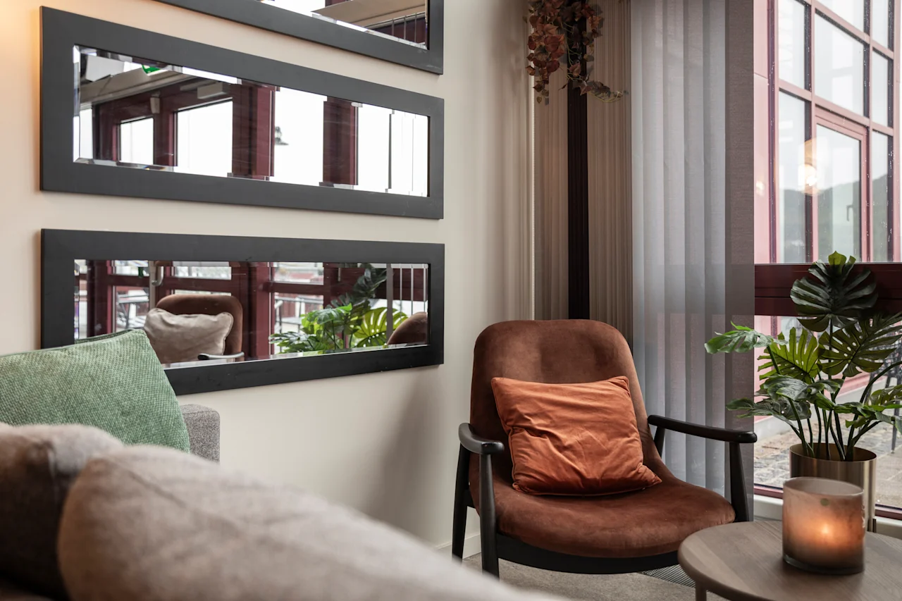 Lämmin ja kutsuva hotellin oleskelutila, jossa on muhkea nojatuoli, pehmeä sohva, tyylikkäät peilit ja luonnonvaloa suuresta ikkunasta.