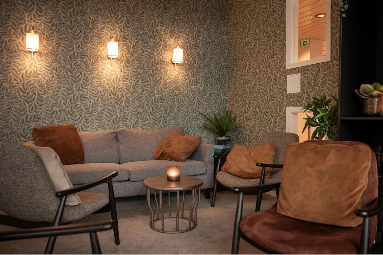 Viihtyisä oleskelutila, jossa on sohva, nojatuoleja ja sytytetty kynttilä sohvapöydällä, lehtikuvioitua tapettia vasten. Poistumisnuoli.