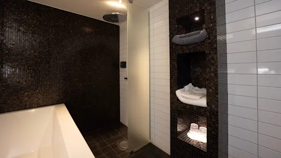 Et moderne badeværelse har et hvidt badekar, en mat glashytte, mørke mosaikfliser og hylder med håndklæder og glas, under klare loftslys.
