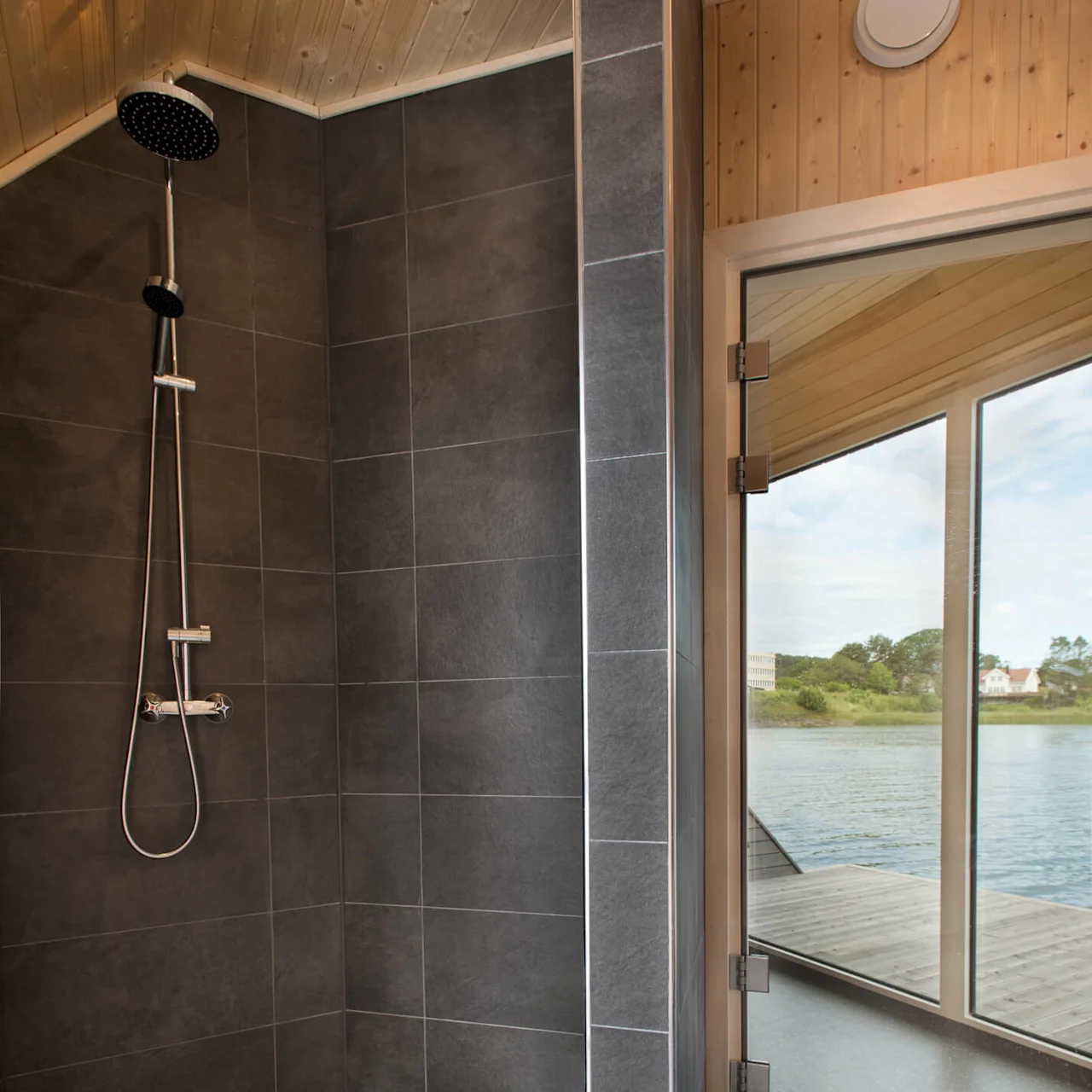 Shower with dark tiles, featuring overhead and handheld fixtures, is adjacent to a glass door. The surroundings include wooden walls and an outdoor water view.