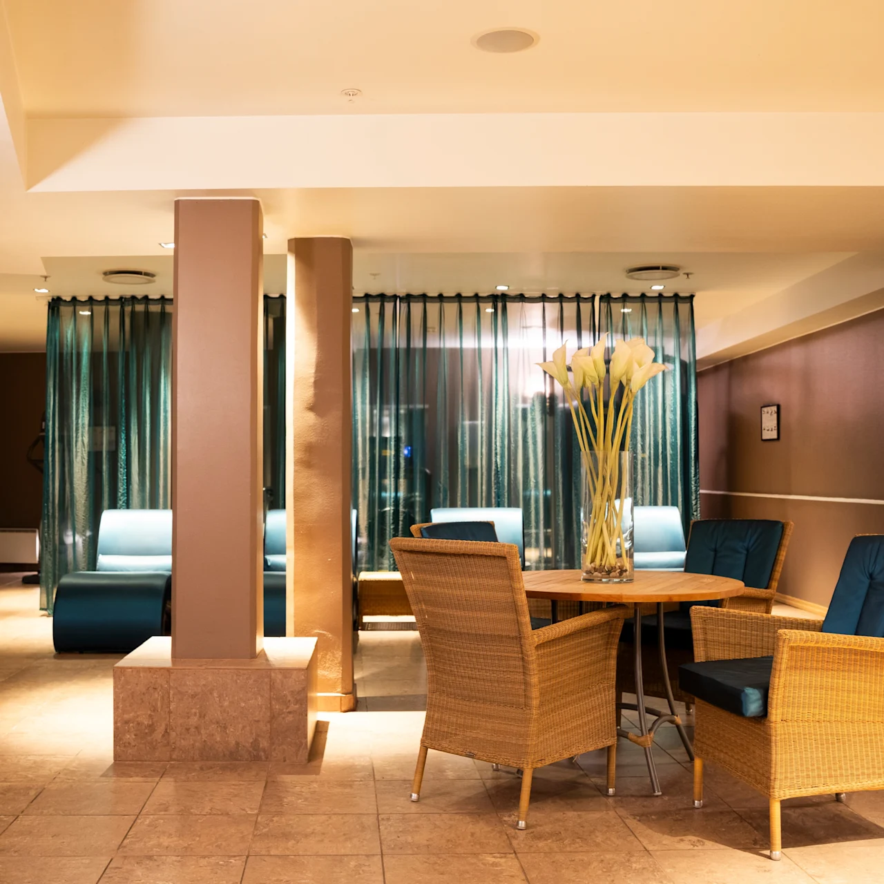 A seating area with wicker chairs surrounds a round table topped with tall decorative grasses, situated in a warmly lit room featuring tiled floors and curtained sections. 