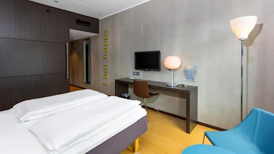 A modern hotel room with a bed, desk, and TV. A chair and lamp are nearby. Wall text reads, "I am happy."