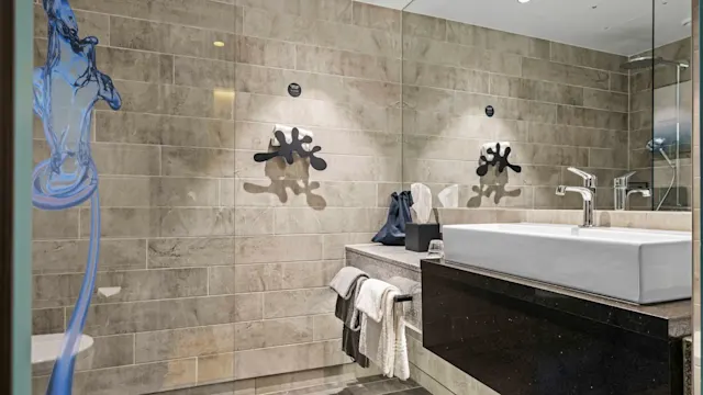 Vask med håndklær hviler på en svart benk; beige flislagt bad med abstrakt kunst på veggene. Glasskillevegg skiller vask fra dusjområde, og skaper et moderne, minimalistisk rom.