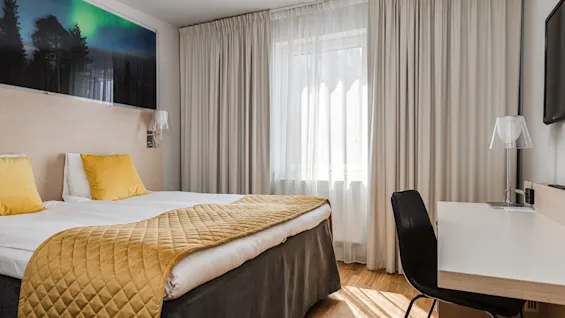 En prydligt bäddad dubbelsäng med gula kuddar och överkast, placerad i ett ljust hotellrum med trägolv, vita gardiner och ett skrivbord med stol nära ett fönster.