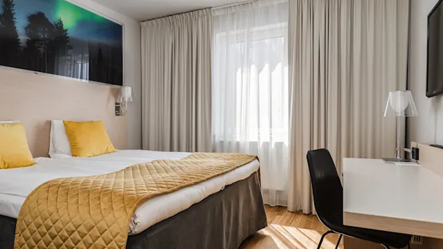 En prydligt bäddad dubbelsäng med gula kuddar och överkast, placerad i ett ljust hotellrum med trägolv, vita gardiner och ett skrivbord med stol nära ett fönster.