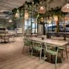 Bord og stoler er arrangert i en stilfull kafé, dekorert med hengende planter og varme pendellamper. Kaféens grønne detaljer og tregulv skaper en hyggelig, innbydende atmosfære.