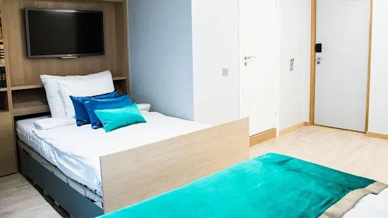 Moderne hotelværelse med en pænt redt seng og turkise puder. Et stort TV er monteret på en træpanel over sengegavlen. Let trægulv og hvide vægge fuldender rummet.