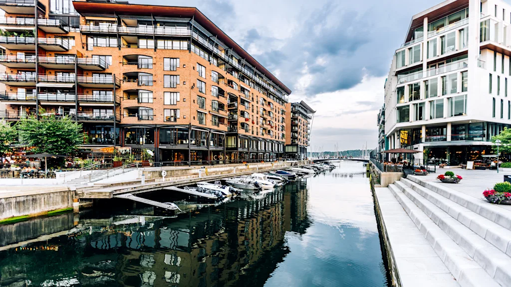 Kanavajohtoiset rakennukset veneineen laiturissa veden äärellä, heijastuen kanavaan. Kohtaus on rajattu moderneilla asunnoilla ja jalankulkukäytävillä, osittain pilvisen taivaan alla.