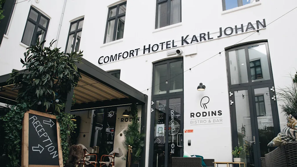 A sign reads "Comfort Hotel Karl Johan" above doors. A chalkboard with plants indicates "Reception" with arrows. Nearby, "Rodins Bistro & Bar" is visible. Tables and chairs are outside. 