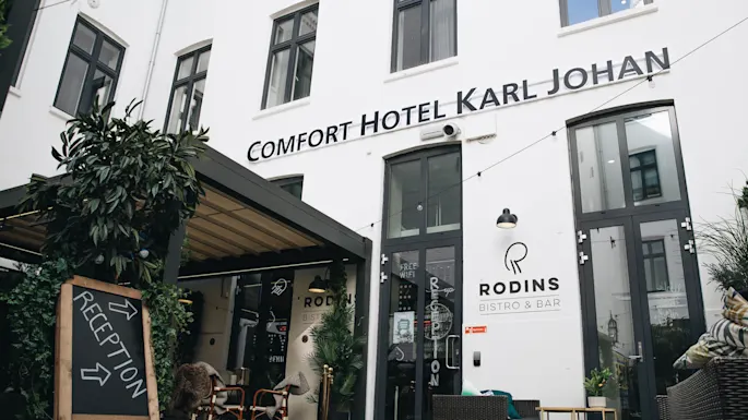 A sign reads "Comfort Hotel Karl Johan" above doors. A chalkboard with plants indicates "Reception" with arrows. Nearby, "Rodins Bistro & Bar" is visible. Tables and chairs are outside. 