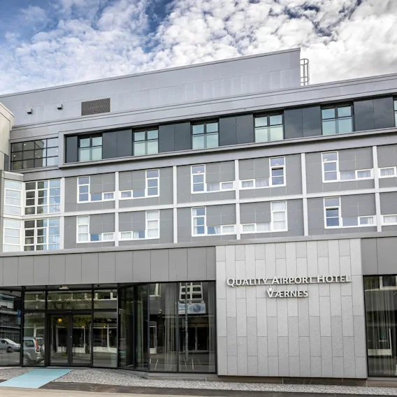 En moderne, grå bygning med skiltet "QUALITY AIRPORT HOTEL VÆRNES" står under en delvis skyet himmel. Store vinduer reflekterer andre strukturer. En mindre "QUALITY HOTEL"-logo er også synlig.