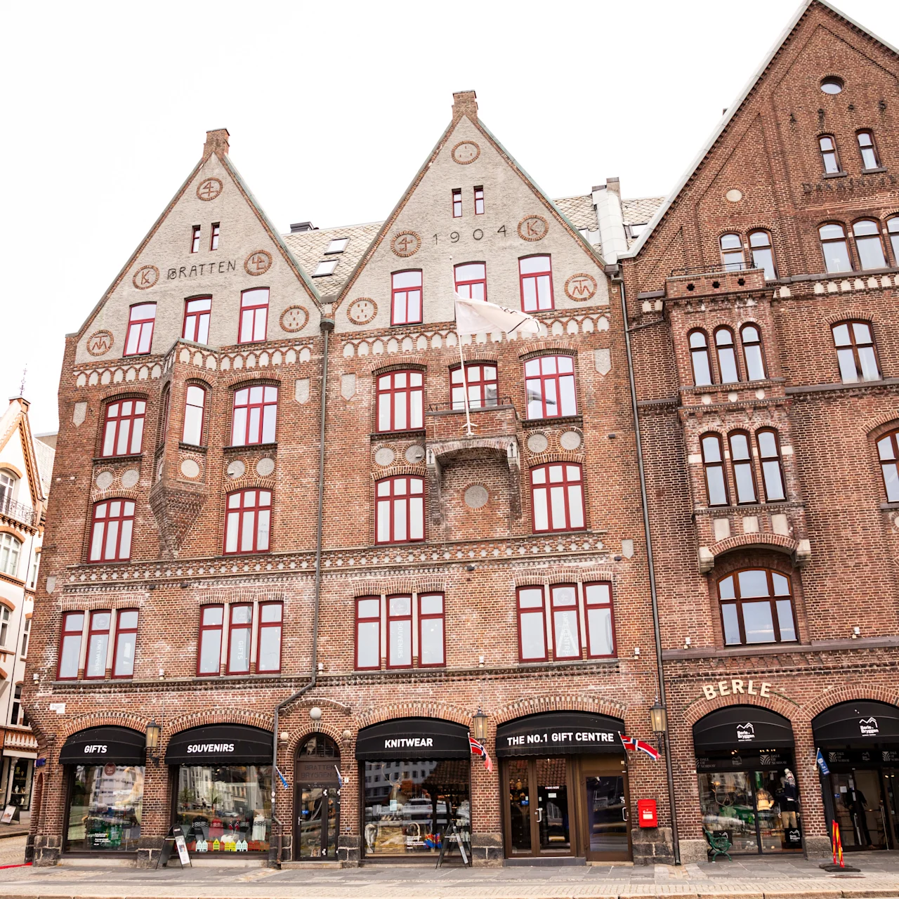 Historisk murstensbygning med gavltage har røde vinduesrammer. Butikker kan ses i form af "GATEAU," "INTERTEKNA," "THE BEST GIFT COMPANY," og "BERLE" på en brostensbelagt gade i bymiljø.