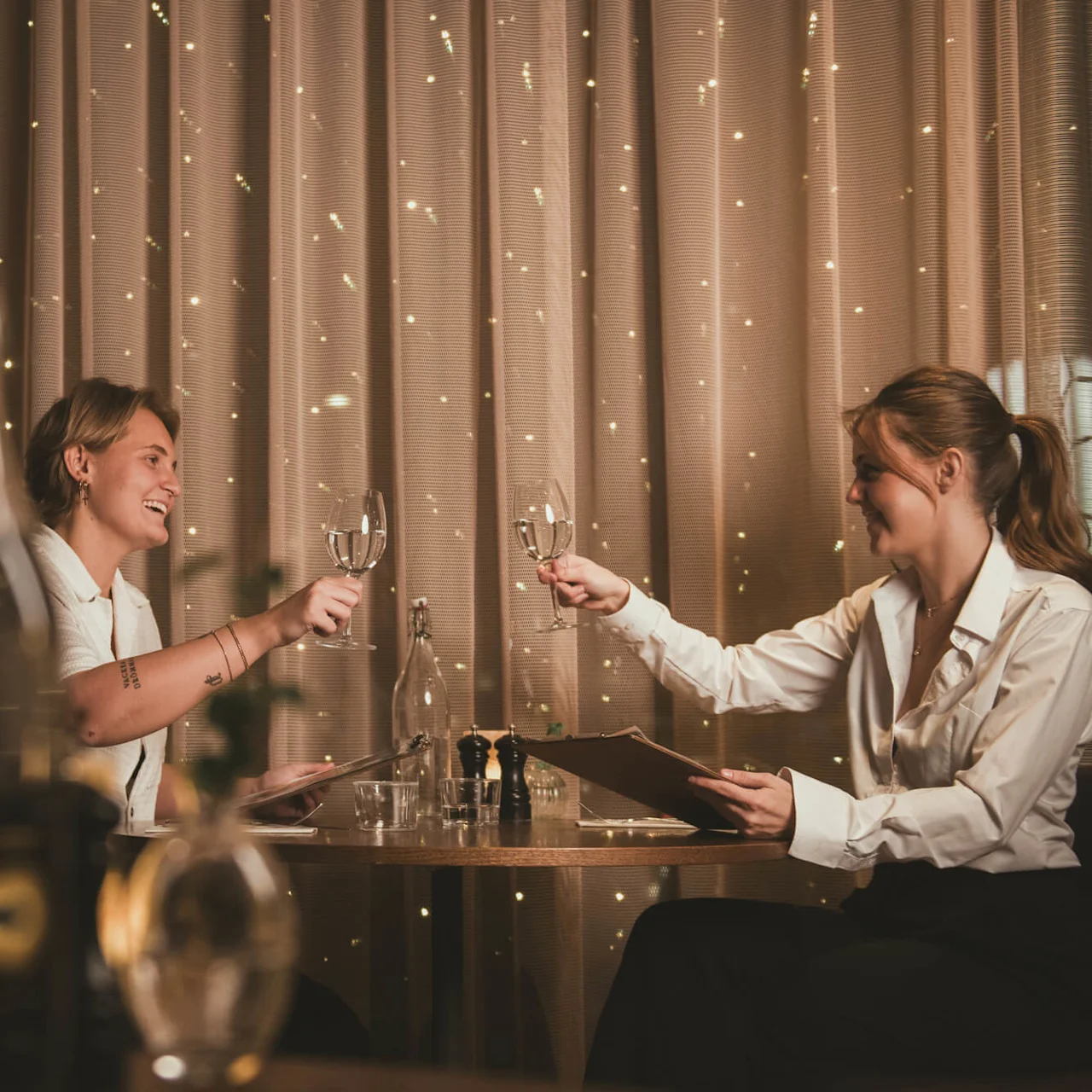 Två personer klingar glas på ett svagt upplyst restaurangbord, leende och hållande menyer. Mjuka ljus glittrar bakom en gardin, vilket skapar en elegant, varm atmosfär.