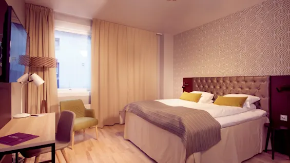 Et pent arrangert hotellrom med mønstret aksentvegg bak en polstret hodegavl, flankert av gardiner og en stol i hjørnet.