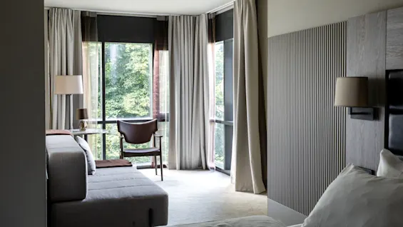 A bright room displays a bed, sofa, and chair beside tall windows with curtains, revealing a green, leafy scene outside.