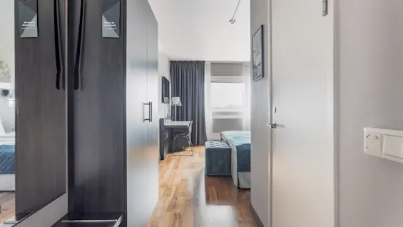 A modern hotel room entrance with a wardrobe, mirror, and 'Do Not Disturb' signs, leading to a bedroom with a bed and window.