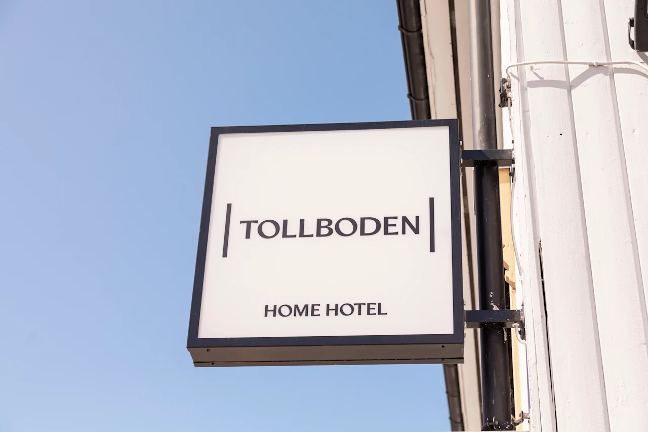 En fyrkantig skylt visar texten "TOLLBODEN" och "HOME HOTEL" i tjocka bokstäver, monterad på en vit byggnad mot en klarblå himmel.