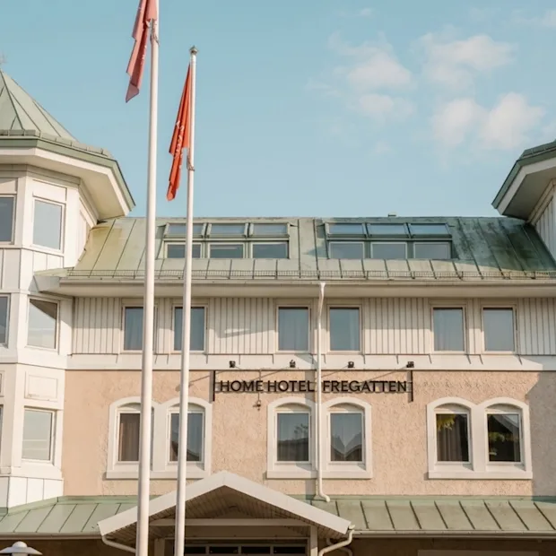 Byggnadens exteriör med en skylt som lyder HOME HOTEL och två flaggor som vajar framför under en klar himmel.