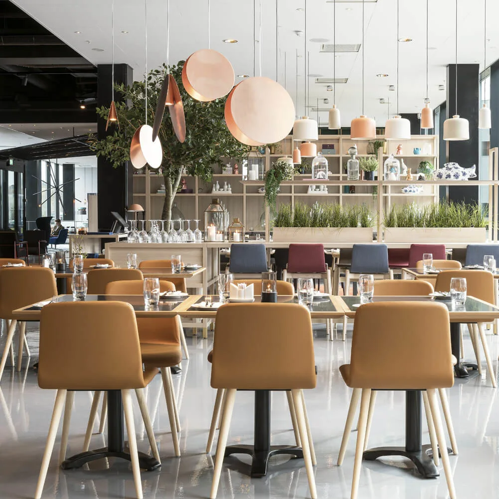 Dining tables with glasses and plates, surrounded by tan chairs, under modern circular pendant lights; set in a bright, contemporary restaurant adorned with potted plants and decorative shelves.