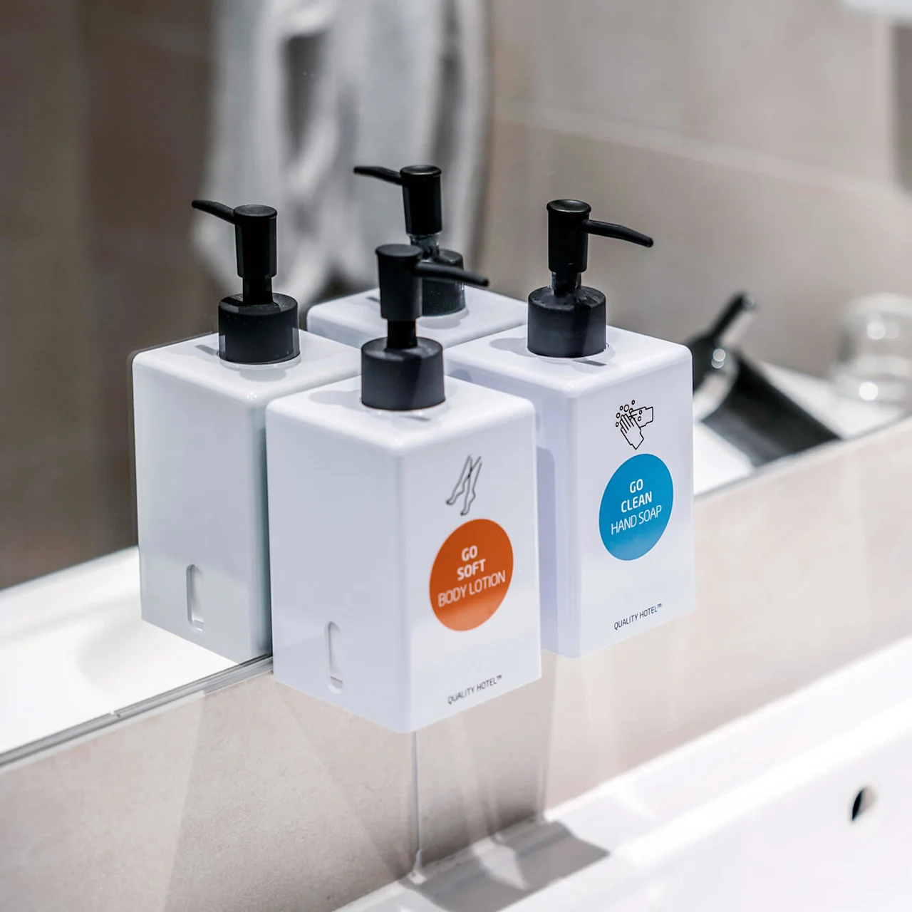 Tre vita dispenserflaskor med svarta pumpar märkta "GO SOFT BODY LOTION" och "GO CLEAN HAND SOAP" är monterade mot en spegel ovanför ett vitt handfat.