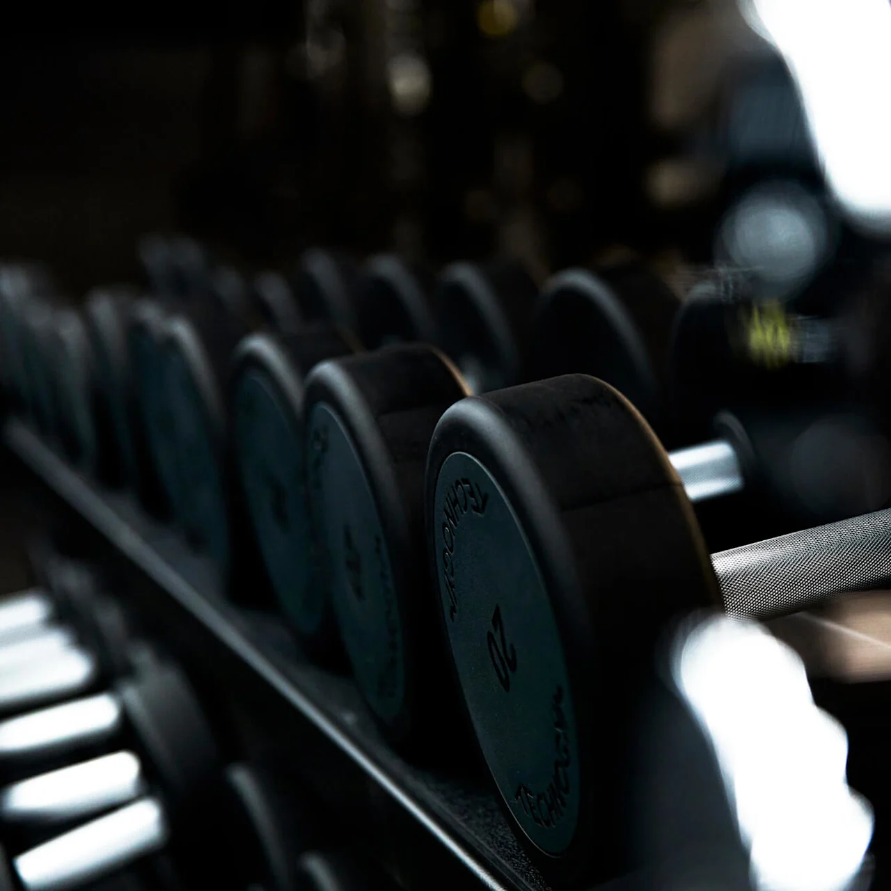 Håndvægte hviler på et stativ i et svagt oplyst træningscenter, fokuseret på en med teksten "TECHNOGYM 20" på siden.