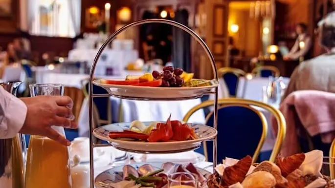 Tiered stand displays fruit, smoked salmon, and cheese; a hand pours orange juice. Elegant dining room with chandeliers and blue chairs in the background.